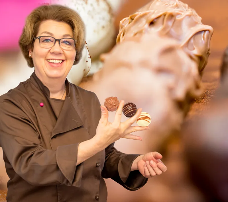 Chocolatiere und Inhaberin Ulrike Maurer hält drei handgemachte Pralinen zwischen den Fingern und lacht. Im Hintergrund ein Close-Up ihres Pralinensortiments.